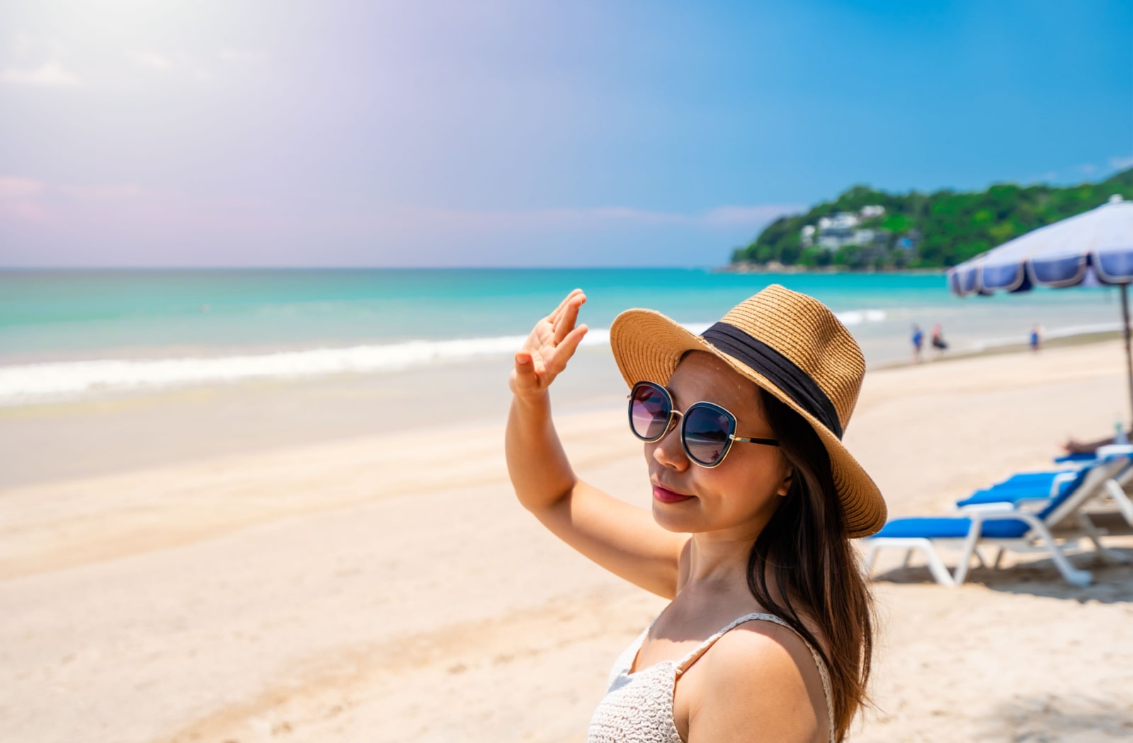 A woman wearing sunglasses, a sun hat, and a bathing suit at the beach, and she's using her hand to block the bright sunlight from her eyes