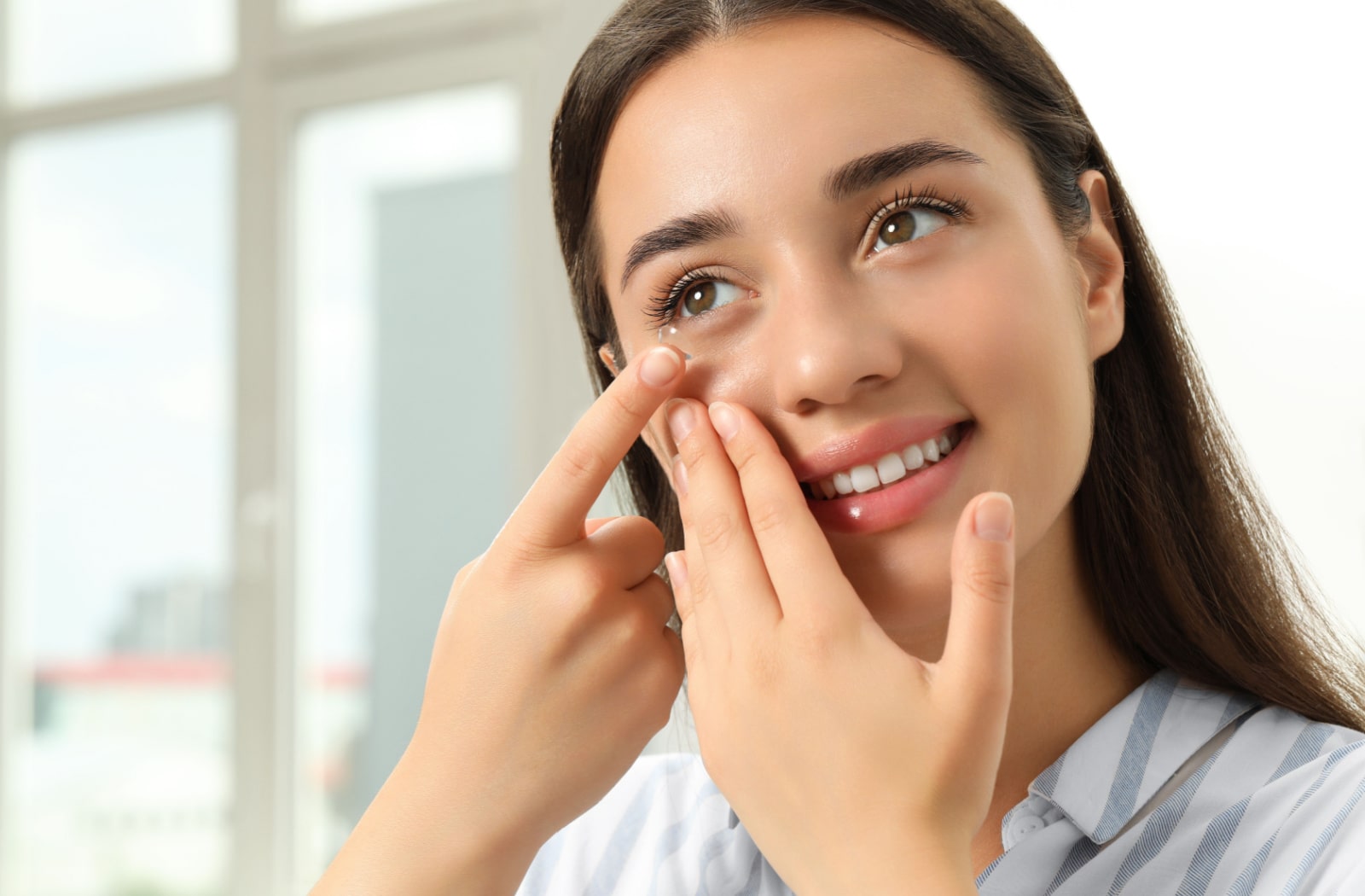 A woman using her right hand to pull her eyelid down while she puts a contact lens on her left eye with her left hand