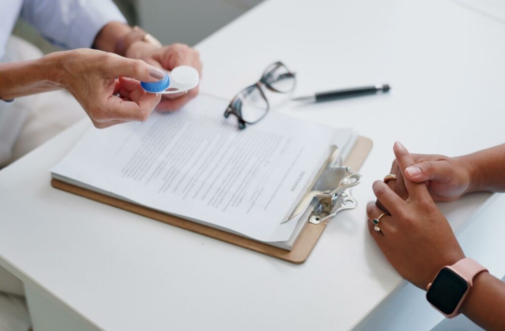 Optometrist holding a contact lens case while discussing lens care with a patient seated across a desk, with paperwork and glasses on the table.