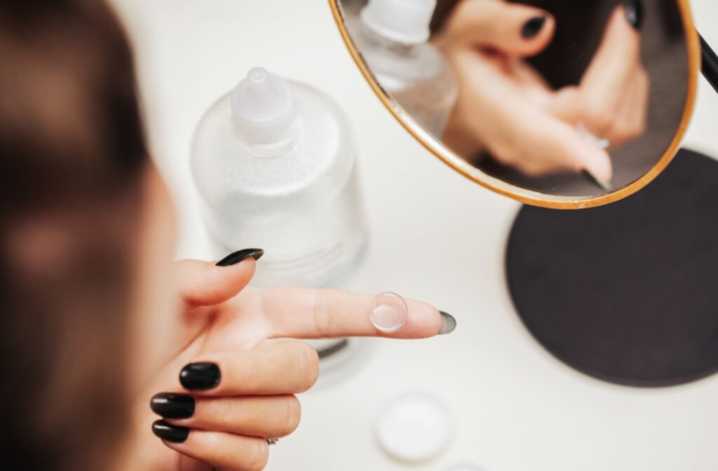 Person with dark nail polish holding a contact lens on their fingertip near a small mirror, with contact lens solution and a lens case on the table.
